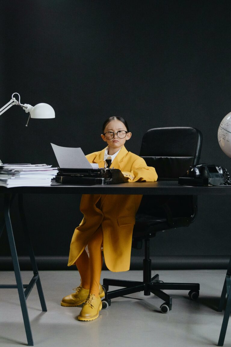 Child in yellow coat and eyeglasses sitting at office desk with typewriter and globe.